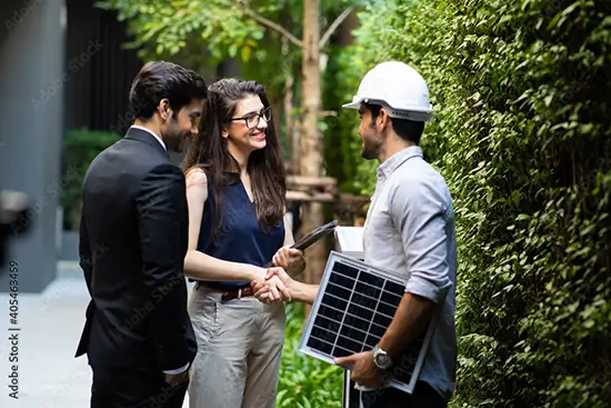 Man holding low wattage solar panel while shaking hands with a woman in group of 3 people outside a building next to a green hedge.
